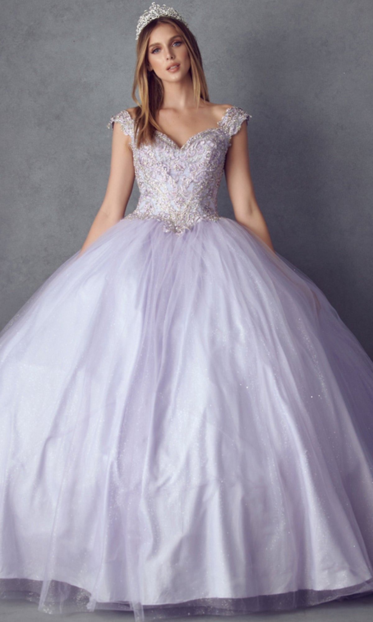 A woman in the Juliet 1430 Cold-Shoulder Glitter Quinceanera Dress, featuring a fitted bodice and off-the-shoulder sleeves, stands against a gray background, dazzling in her lavender gown and tiara.